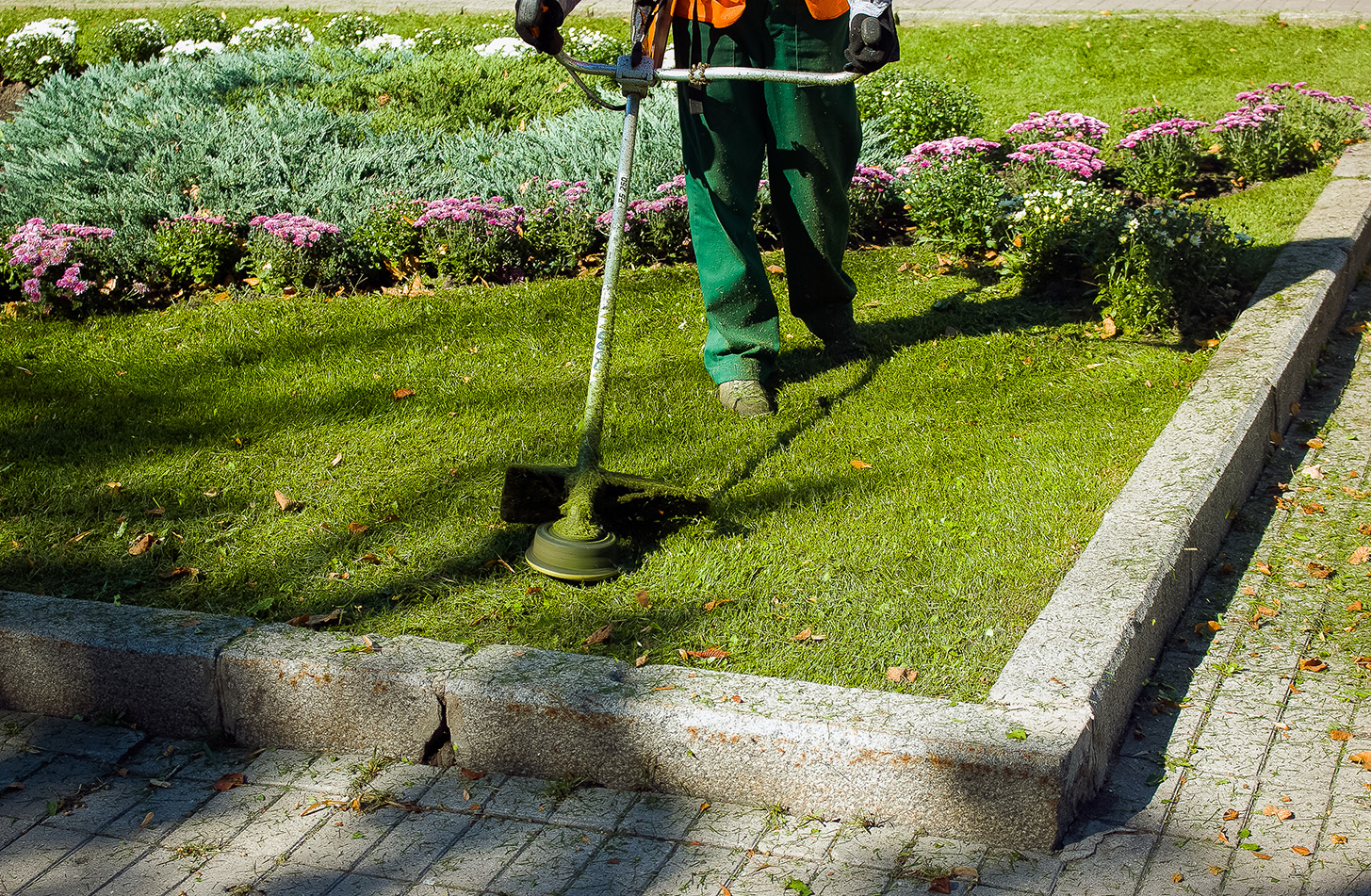 A person wearing an orange safety vest and green pants uses a string trimmer to cut grass along the edge of a paved walkway. To the left is a garden bed with various plants, including some with purple flowers.