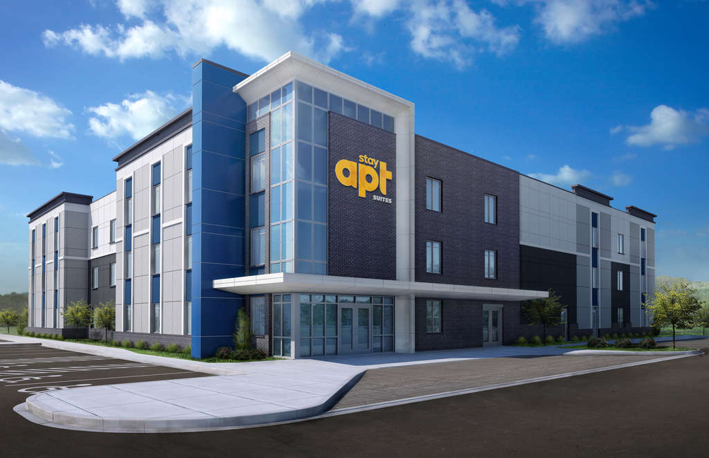 Modern three-story StayAPT Suites hotel building with glass entrance and blue and gray exterior under a partly cloudy sky.