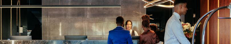 Hotel reception with a female receptionist assisting two guests at the front desk while a bellhop walks by.
