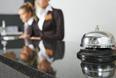 Reception desk with a service bell in focus and two staff members blurred in the background.