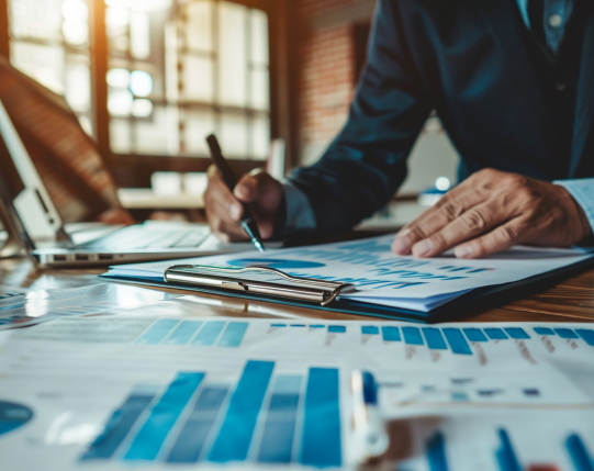 Person in business attire reviewing and marking charts and graphs on paper at a desk with a laptop nearby.