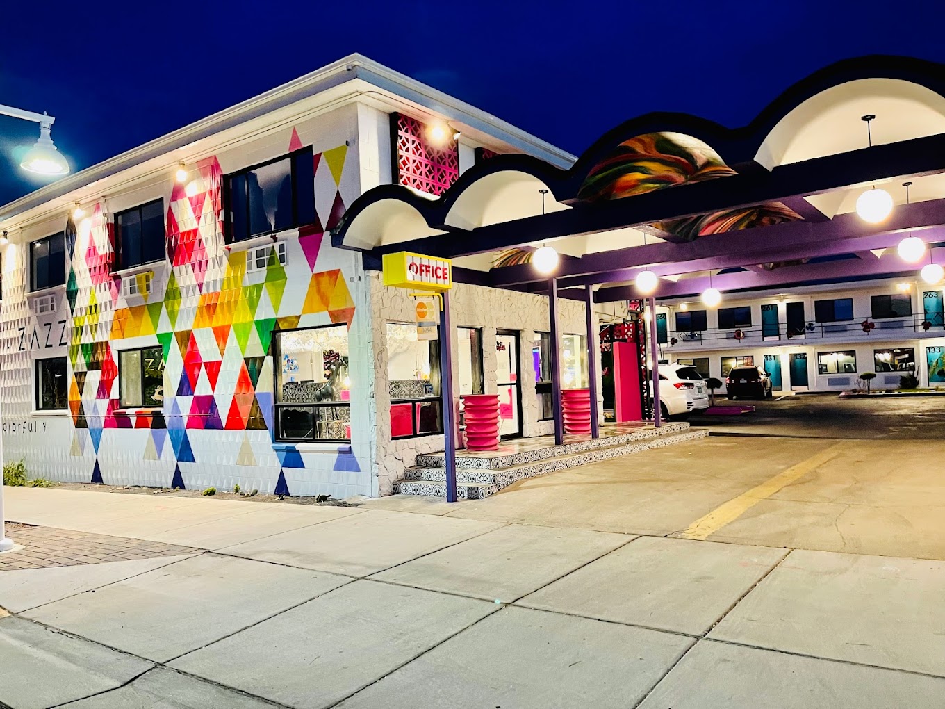 Colorful motel exterior at night with a vibrant geometric mural and a lit office entrance under curved canopies.