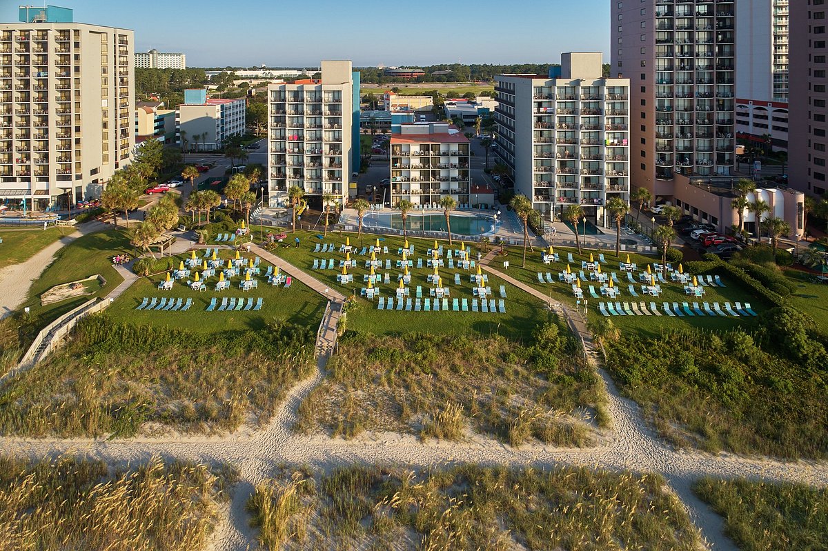 Aerial view of a beachfront resort with lawn chairs, umbrellas, palm trees, and high-rise buildings near a sandy beach path.