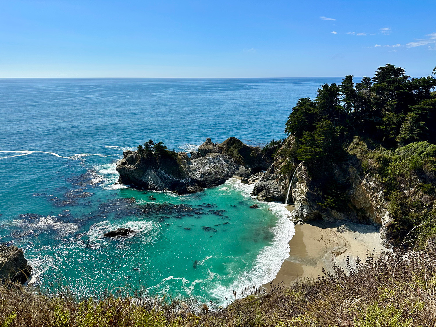A beach in Big Sur, California sits below a cliffside. Bluegreen waves crash against the shore and are framed by a lush green forest that grows right up to the edge of the cliffs.