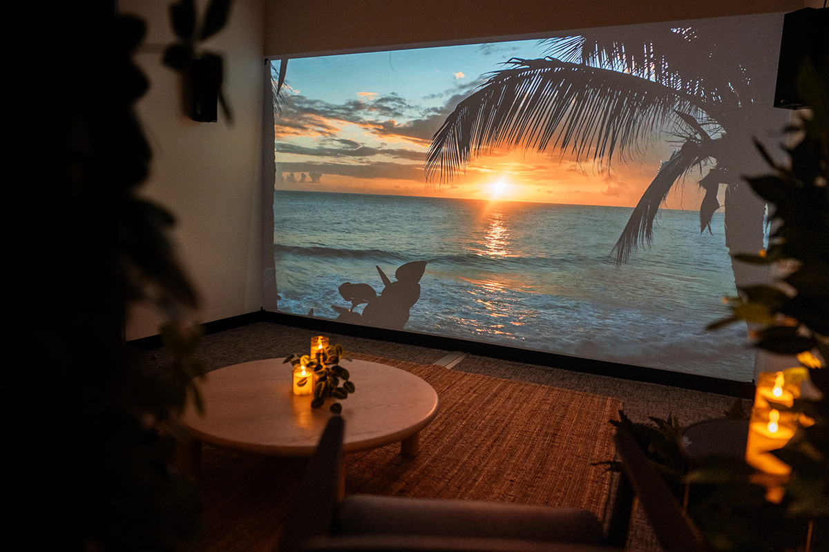 A room inside of an office building sits with candles glowing, a projection on the wall of an Applied Wonder experience, an alcove of plants, sitting chairs, and other decor such as a coffee table and side table.