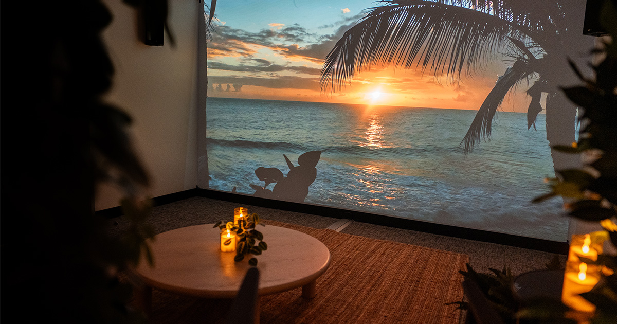 A room inside of an office building sits with candles glowing, a projection on the wall of an Applied Wonder experience, an alcove of plants, sitting chairs, and other decor such as a coffee table and side table.