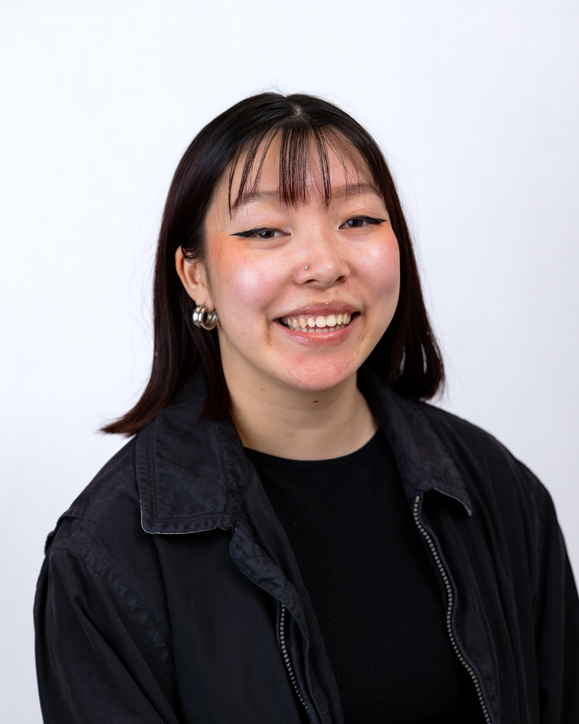 A Southeastern Asian individual with dark brunette hair and light skin. They have bangs and shoulder-length hair. They are wearing small, chunky silver hoops and is dressed in a black t-shirt and jacket. 