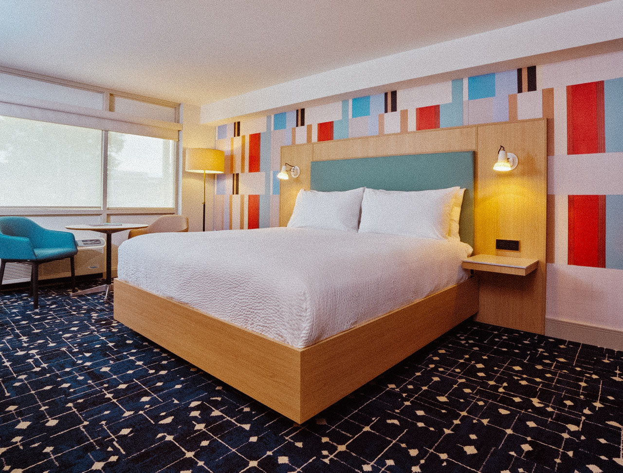 A picture of a room at CHML, a well lit room with a colorful wall pattern and a geometric floor pattern with a queen bed in the center.