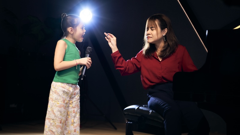 A young student (green) is singing her heart out as her teacher (red) conducts her pitch with her hands.