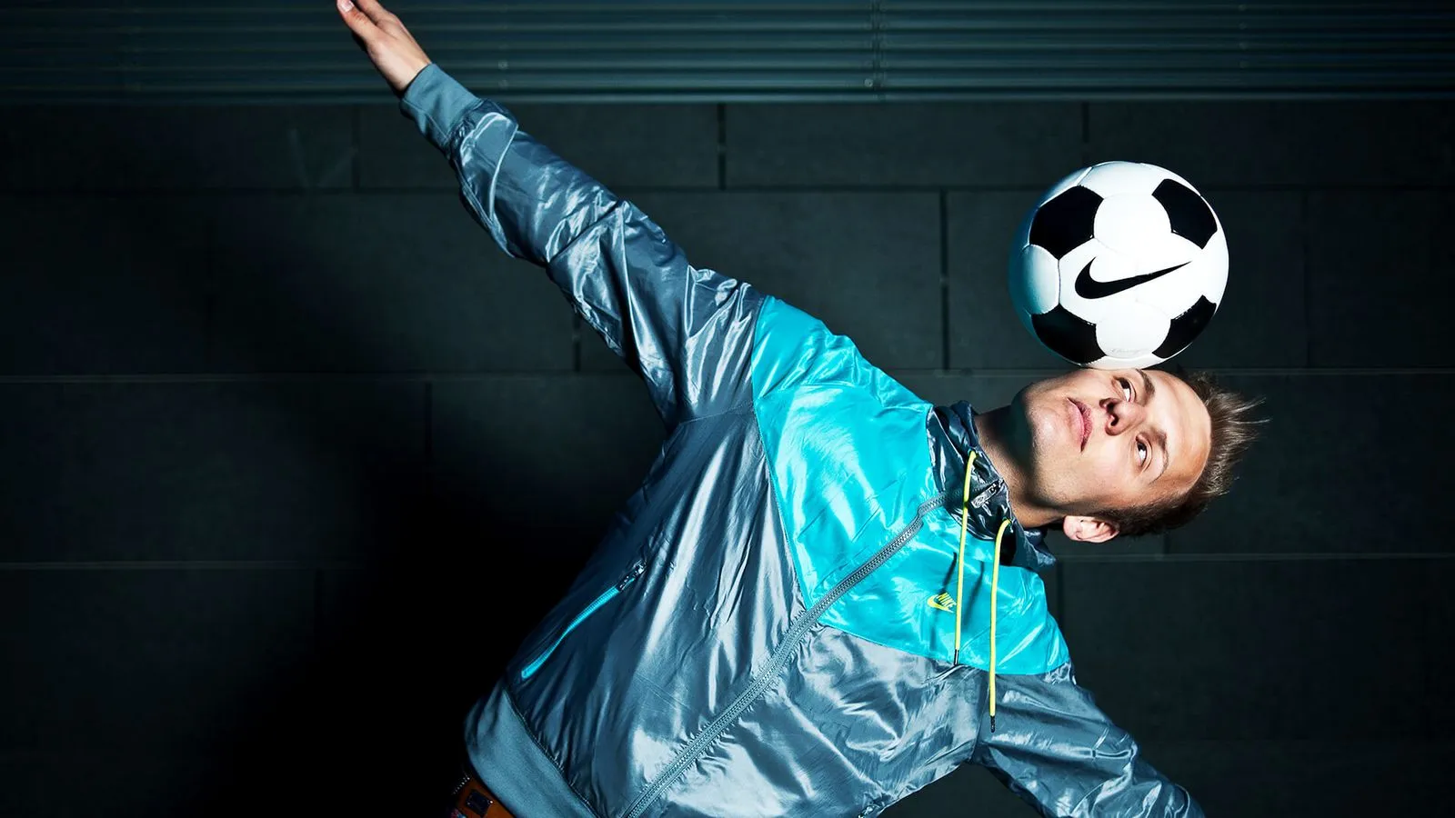 Mann balanciert einen Fußball mit Nike-Logo auf seiner Stirn, trägt einen blauen Trainingsanzug.