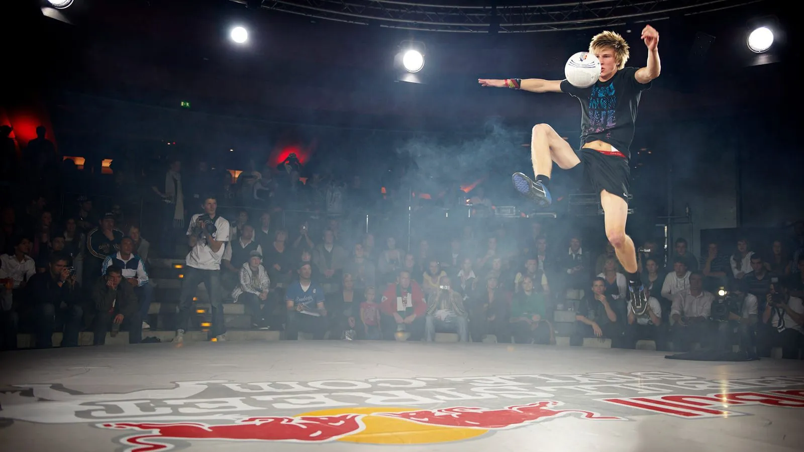 Junger Mann in schwarzer Sportkleidung führt einen Trick mit einem Fußball auf einer Bühne mit Red Bull Logo vor, umgeben von zuschauendem Publikum.
