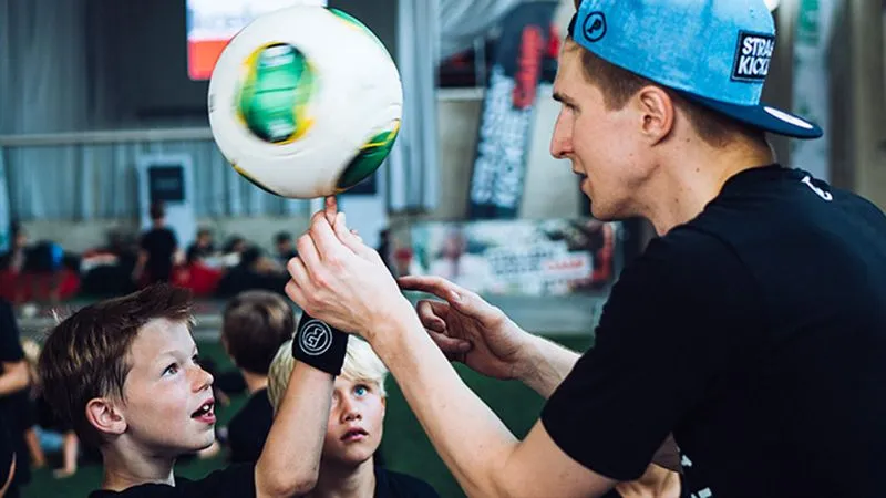 Ein Mann in blauer Kappe zeigt zwei Jungen, wie man einen Fußball auf dem Finger balanciert.