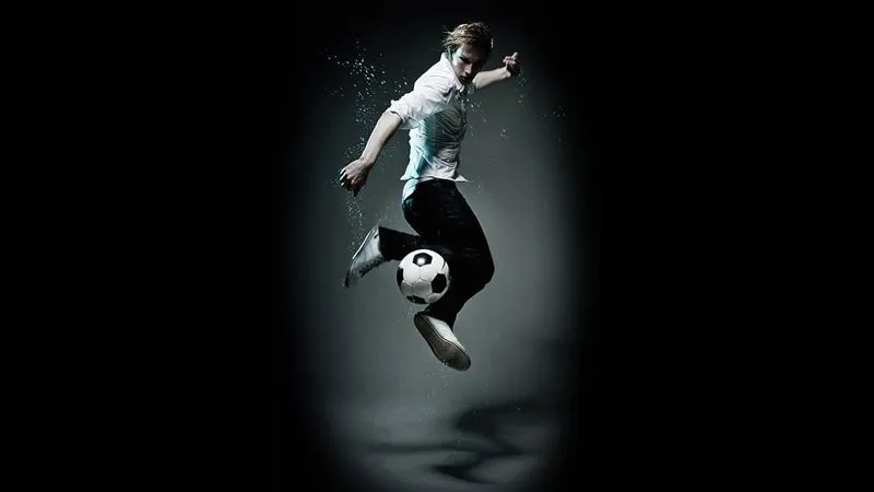 Junger Mann springt hoch und spielt Fußball mit einem Ball vor dunklem Hintergrund.