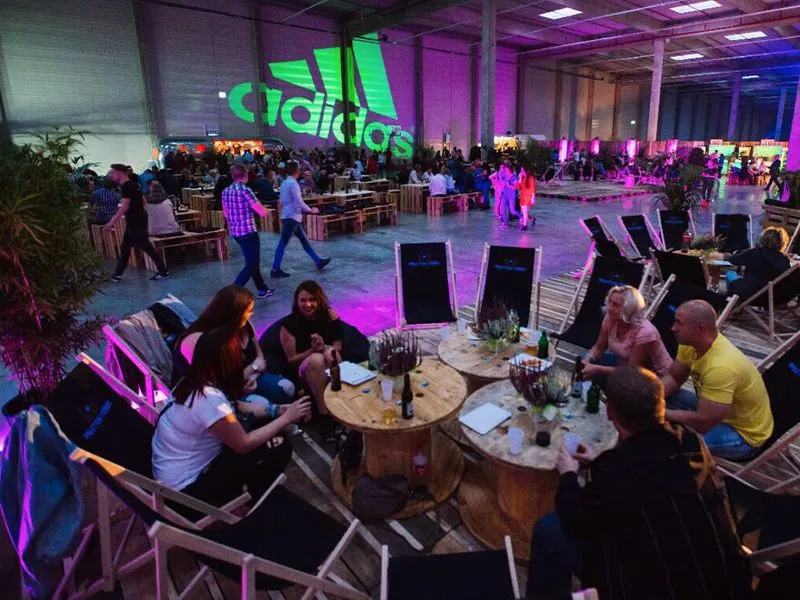 Menschen sitzen auf Liegestühlen um niedrige runde Holztische in einer großen Halle mit Adidas-Logo an der Wand und stimmungsvoller bunter Beleuchtung.