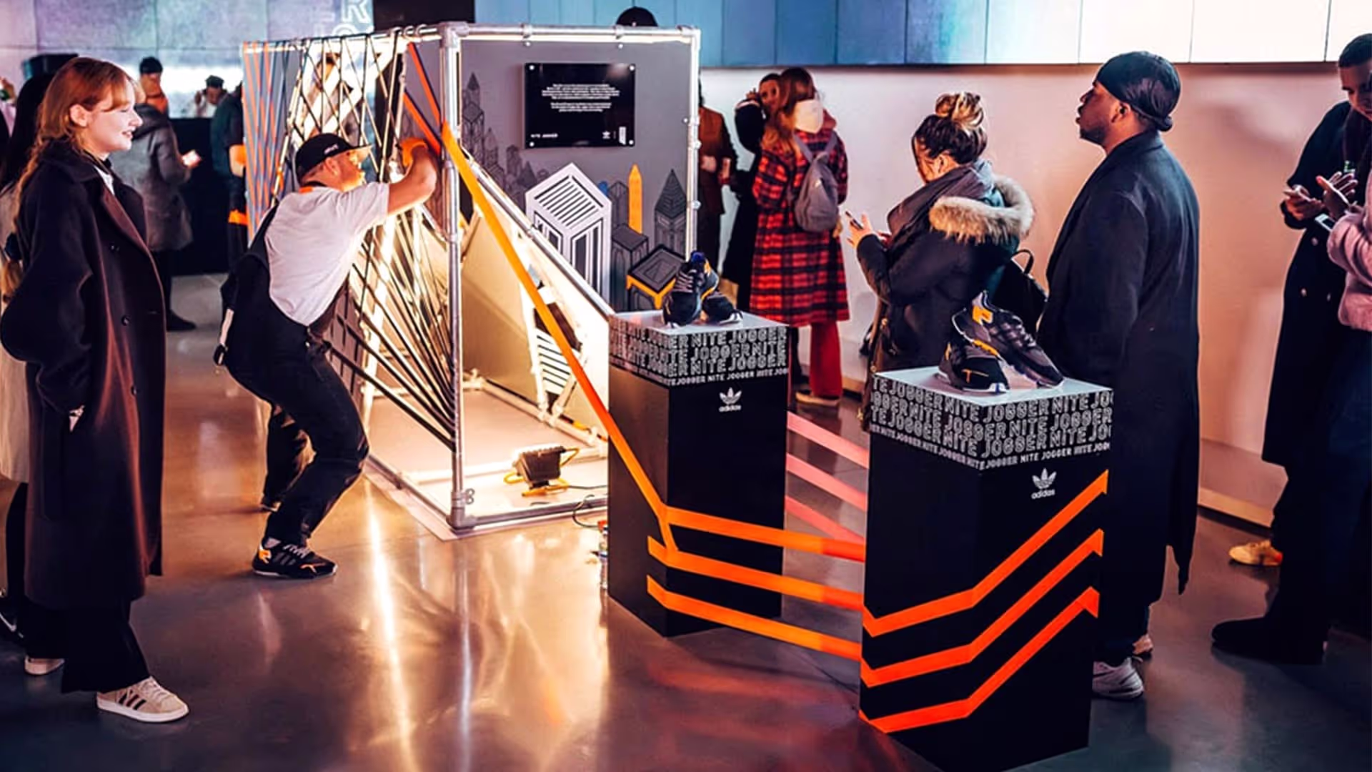Menschen bei einer Markenpräsentation mit Adidas Schuhen auf Podesten und orangefarbenen Bändern, die eine interaktive Installation bilden.