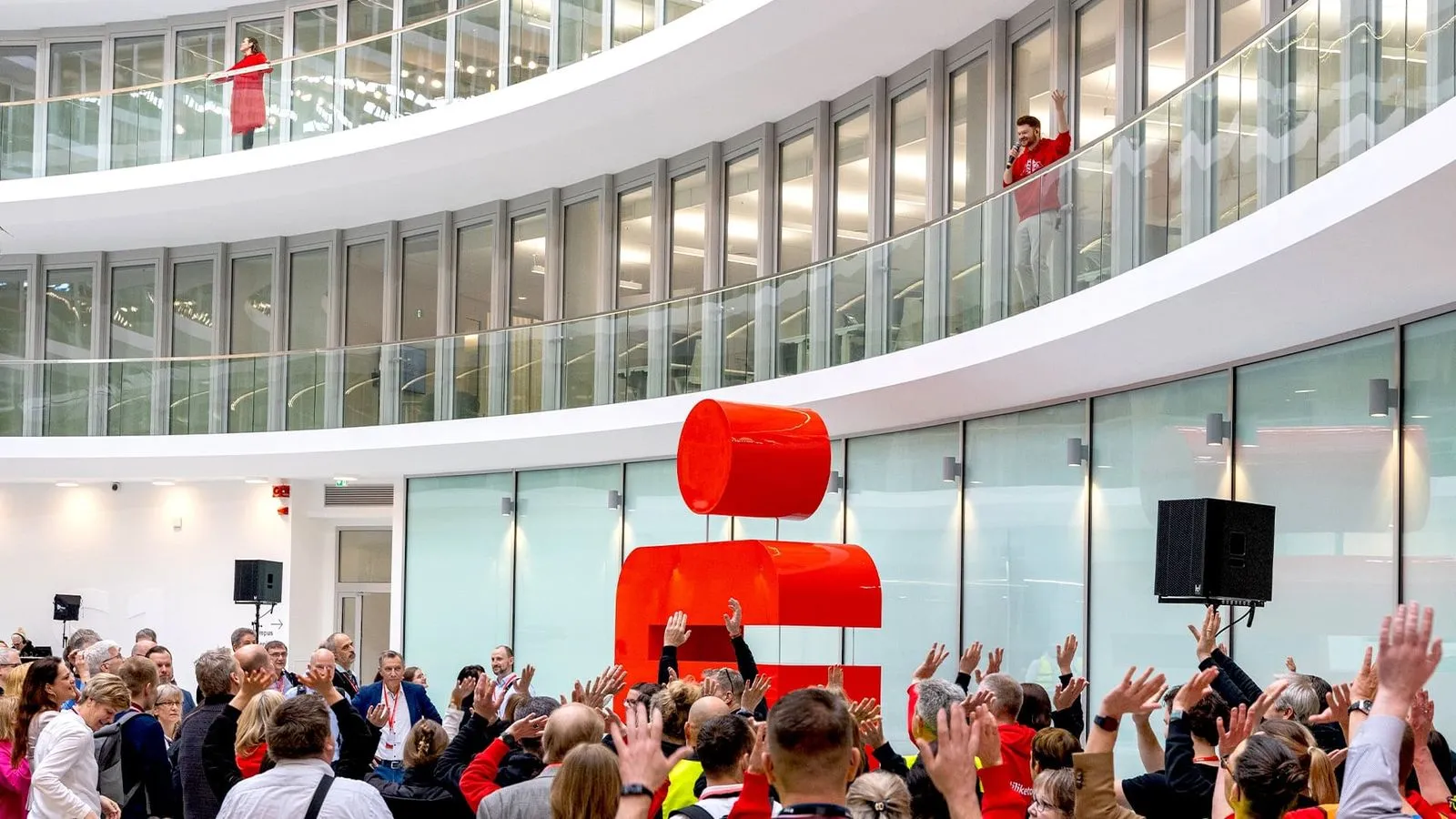 Menschenmenge hebt Hände vor großer roter Skulptur in modernem Gebäude mit Glasgalerien.