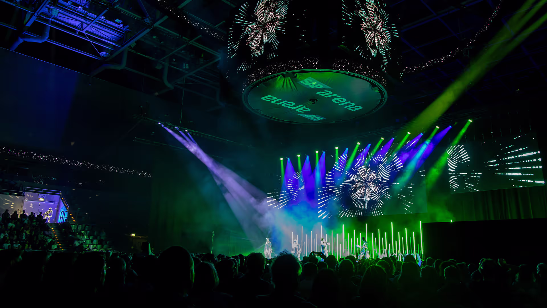 Bühnenperformance mit grünen und violetten Scheinwerfern, digitaler Hintergrundprojektion und Publikum in der SAP Arena.
