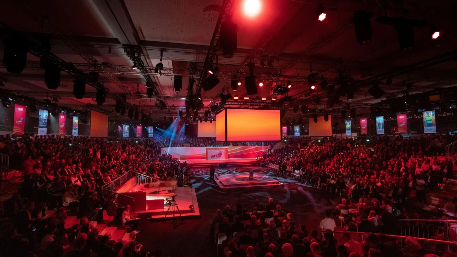 Große Veranstaltungshalle mit roter Beleuchtung, voll besetztem Publikum und zentraler Bühne mit großen Bildschirmen.