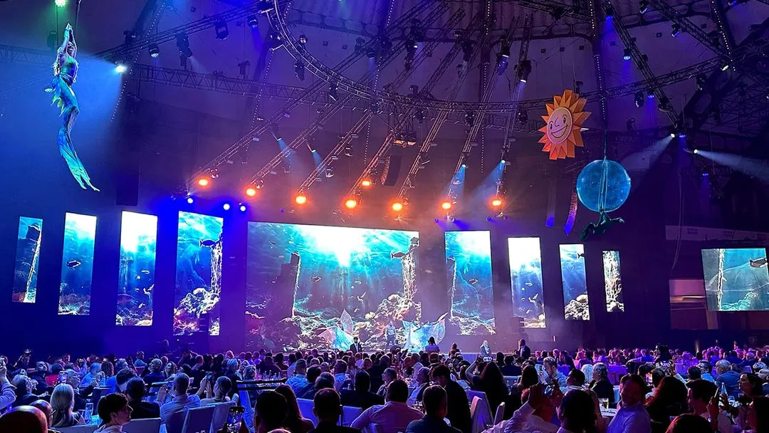 Große Menschenmenge sitzt vor einer Bühne mit mehreren Bildschirmen, die ein Unterwasserbild zeigen, und eine Person erscheint an einem Seil in der Luft.
