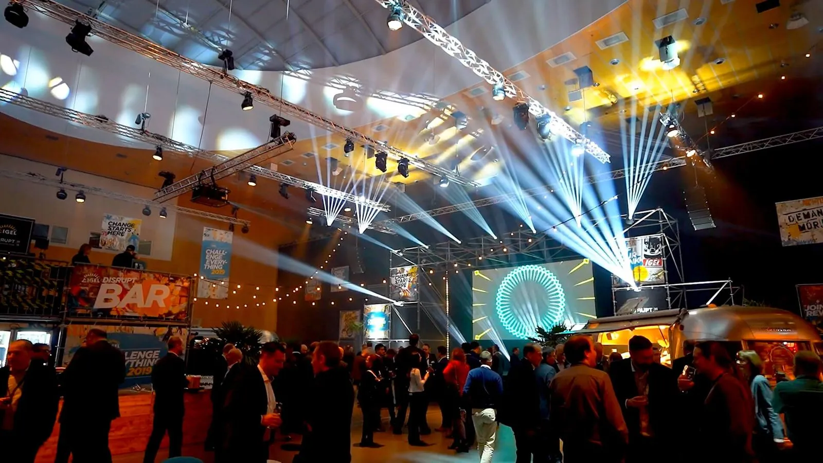 Menschen versammeln sich in einem Veranstaltungsraum mit Bar, Bühnenbeleuchtung und großer Leinwand mit grafischem Muster.