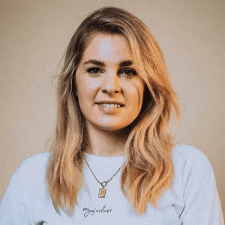 Porträt einer jungen Frau mit langen blonden Haaren, die ein weißes Oberteil und eine Halskette mit einem Anhänger trägt.