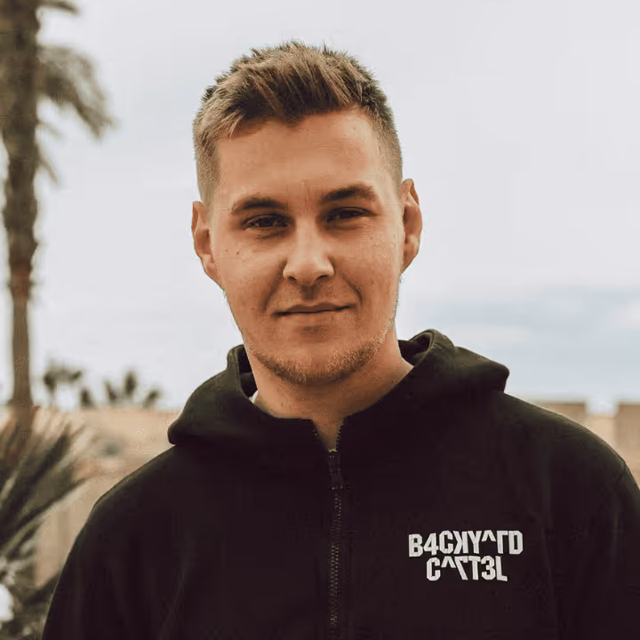 Junger Mann mit kurzem blonden Haar trägt schwarzen Kapuzenpullover mit 'BACKYARD CATS' Schriftzug, unscharfer Außenhintergrund mit Palmen.