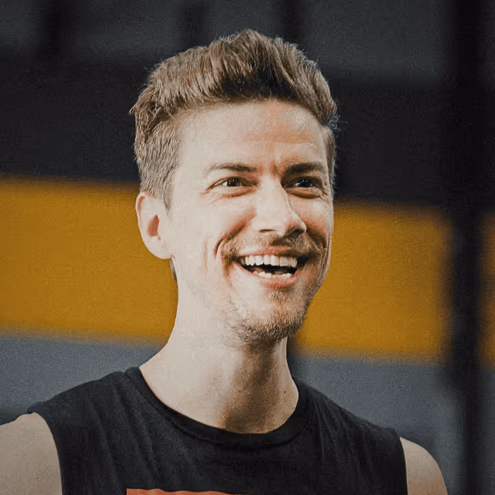 Lächelnder junger Mann mit braunen Haaren und kurzem Bart vor unscharfem Hintergrund mit gelbem und grauem Bereich.