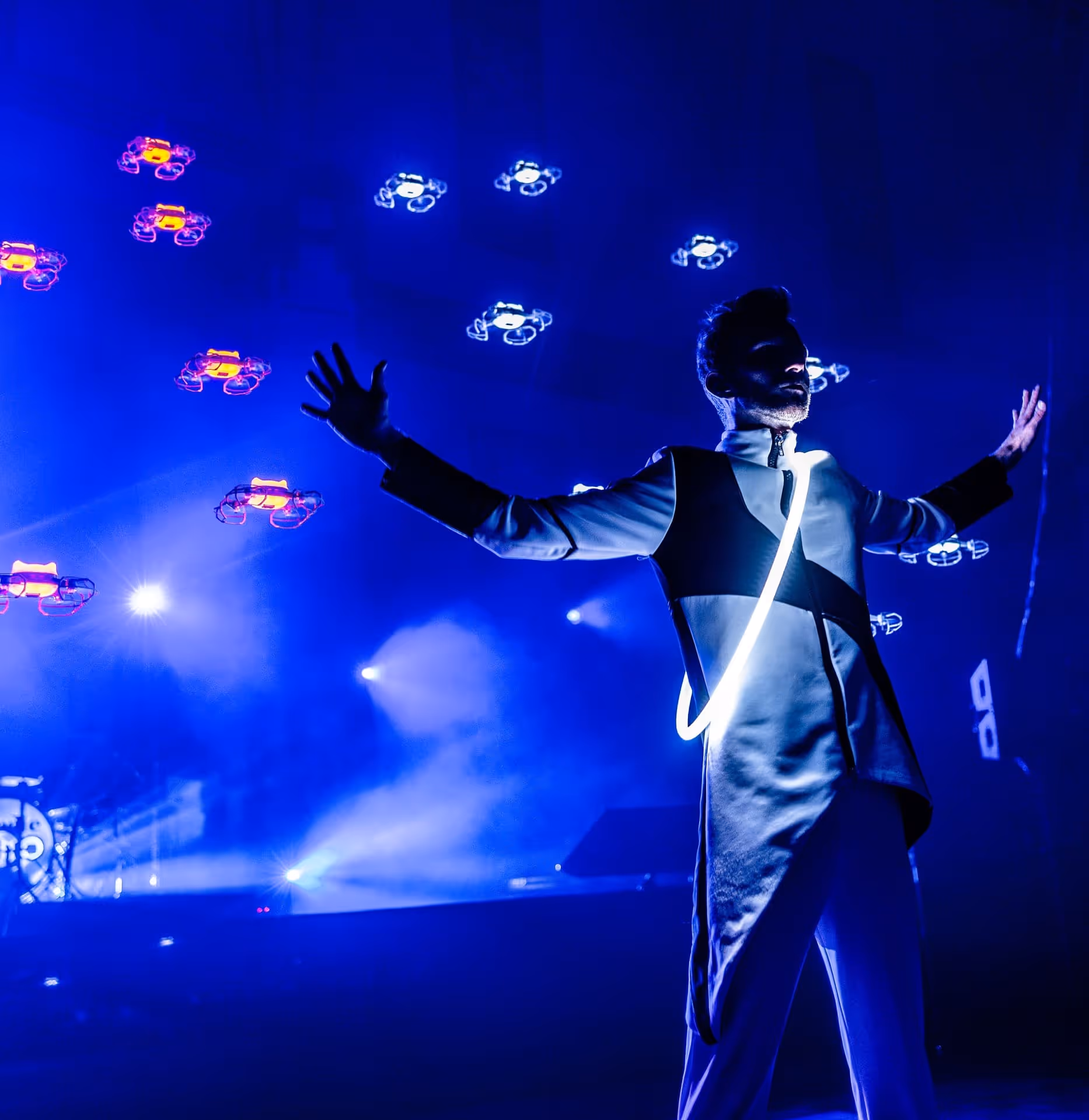 Ein Mann in futuristischer Kleidung mit leuchtendem Band steht mit ausgebreiteten Armen vor einem dunklen Hintergrund mit fliegenden Drohnen und blauer Bühnenbeleuchtung.
