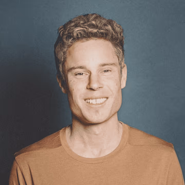 Junger Mann mit lockigem Haar lächelt in die Kamera vor blauem Hintergrund.