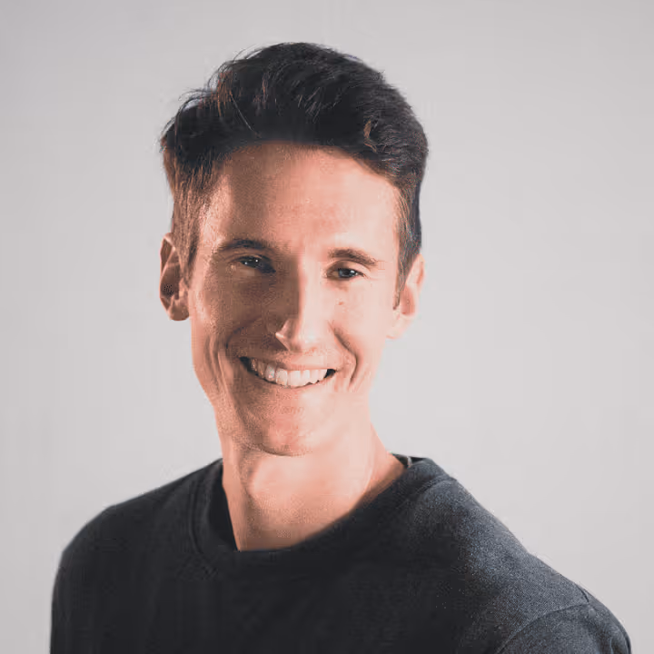 Junger Mann mit kurzem braunem Haar und schwarzem Pullover lächelt in die Kamera vor hellem Hintergrund.