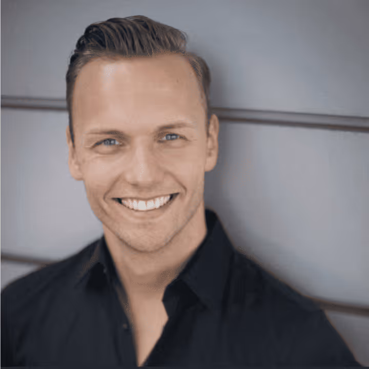 Lächelnder Mann mit kurz zurückgekämmtem blonden Haar und schwarzem Hemd vor grauem Hintergrund.