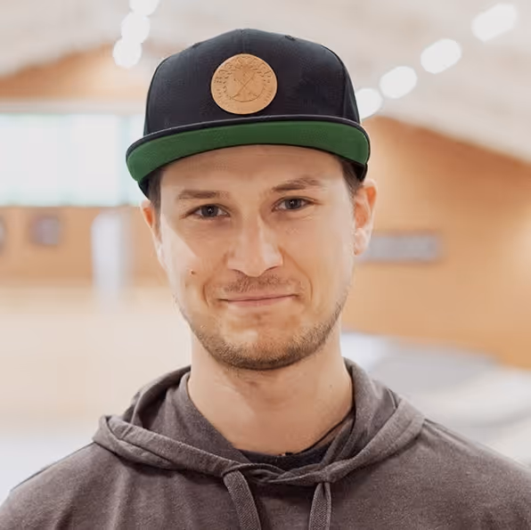 Porträt eines jungen Mannes mit dunkler Kappe und grauer Kapuzenjacke vor unscharfem Skatepark-Hintergrund.