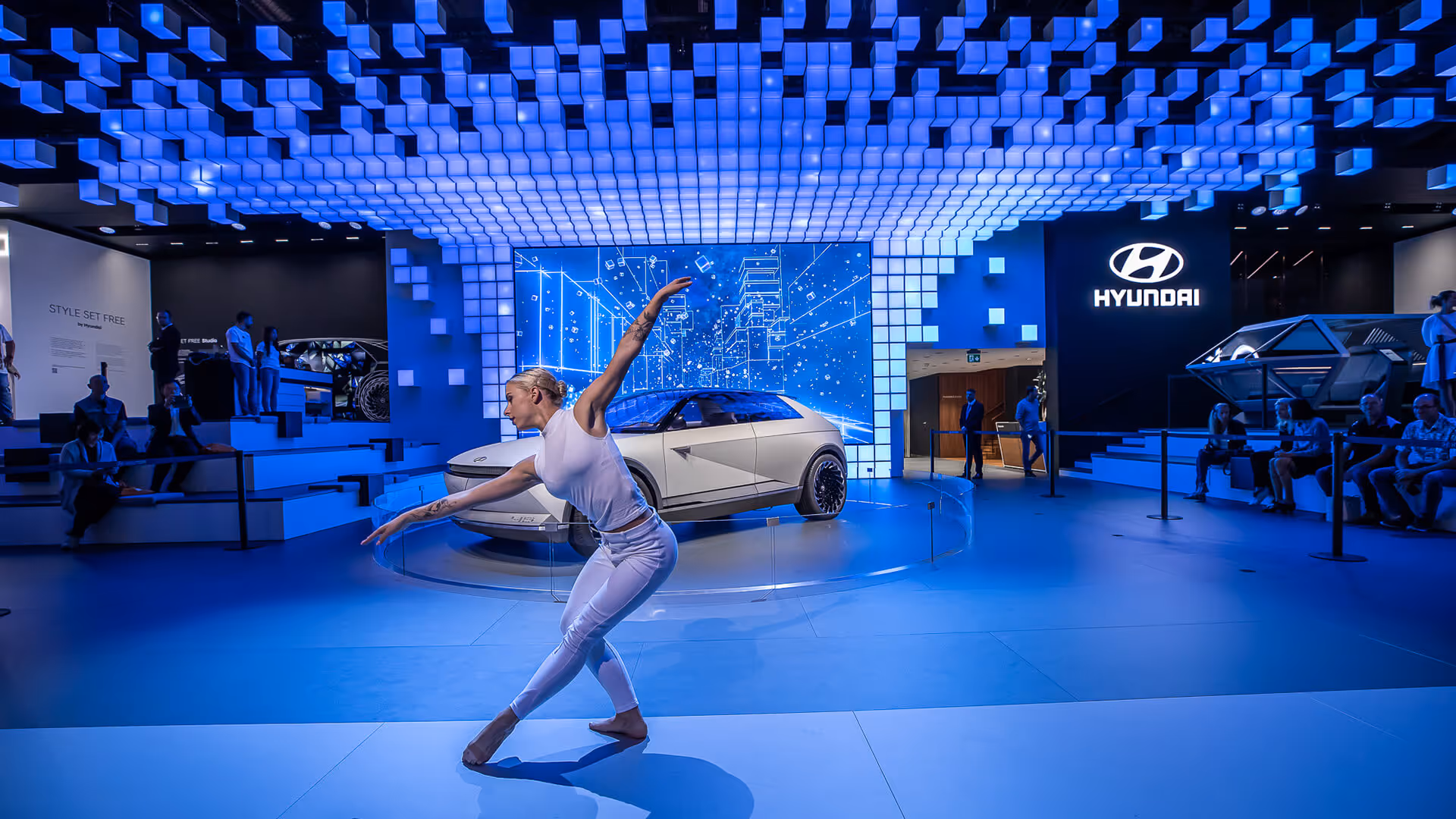 Tänzerin in weißer Kleidung führt eine elegante Pose vor einem futuristischen Hyundai-Auto und einer blauen pixelartigen Deckenbeleuchtung aus.