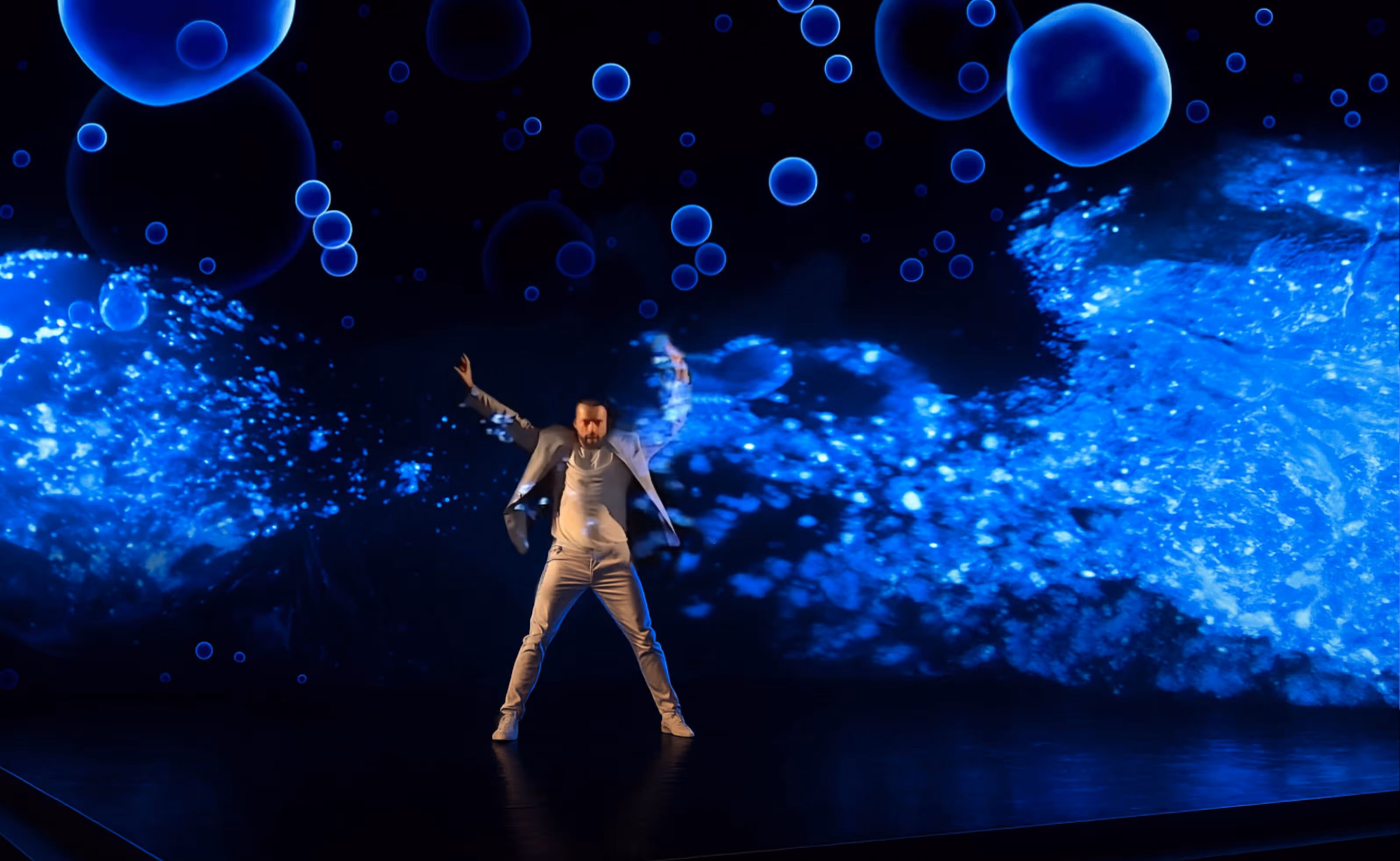 Ein Mann in weißem Anzug tanzt vor einer großen Leinwand mit leuchtenden blauen Blasen und abstrakten Mustern.