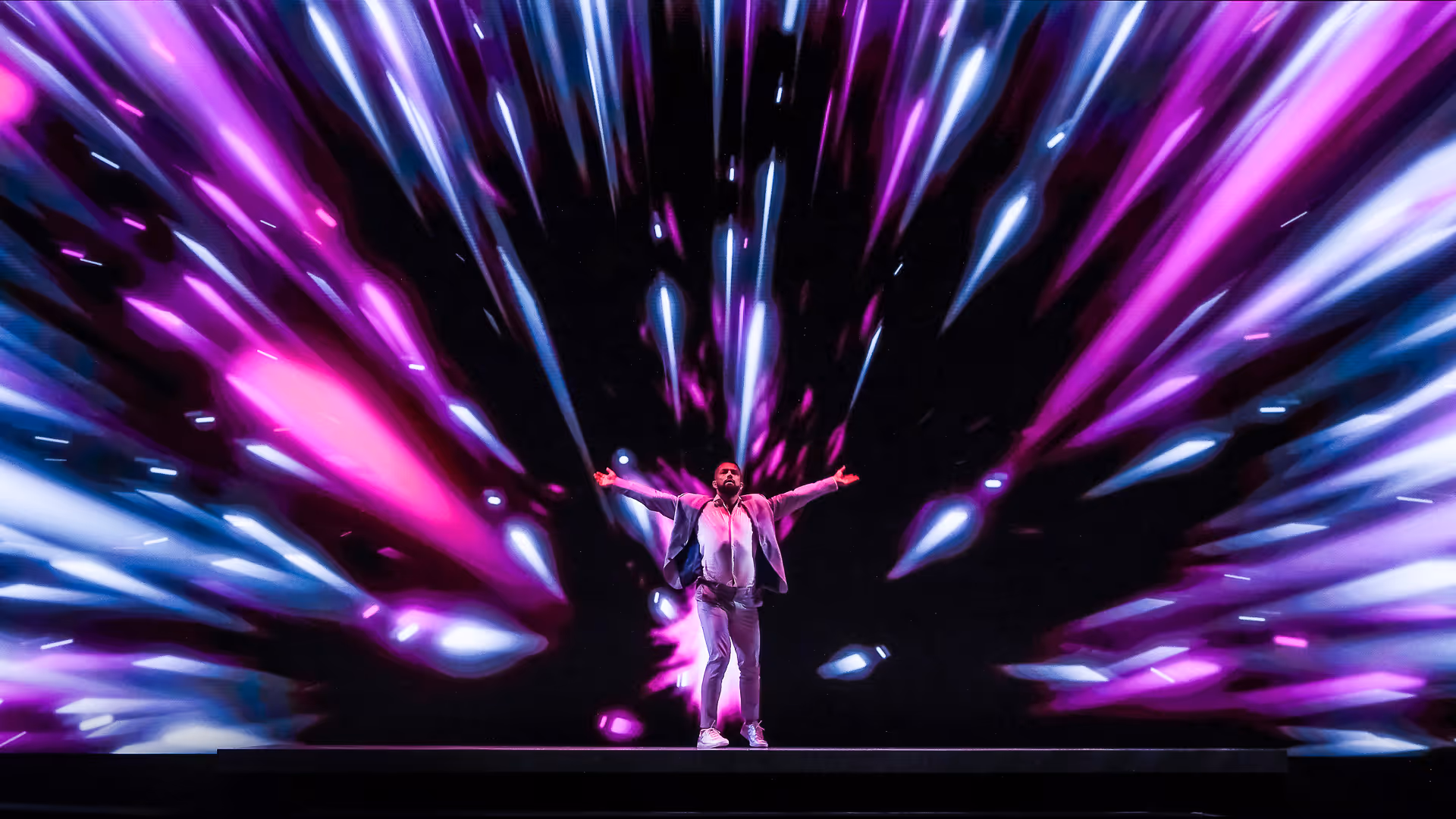 Ein Mann auf einer Bühne steht mit ausgestreckten Armen vor einem lebhaften Hintergrund aus pinken und weißen Lichtstrahlen.