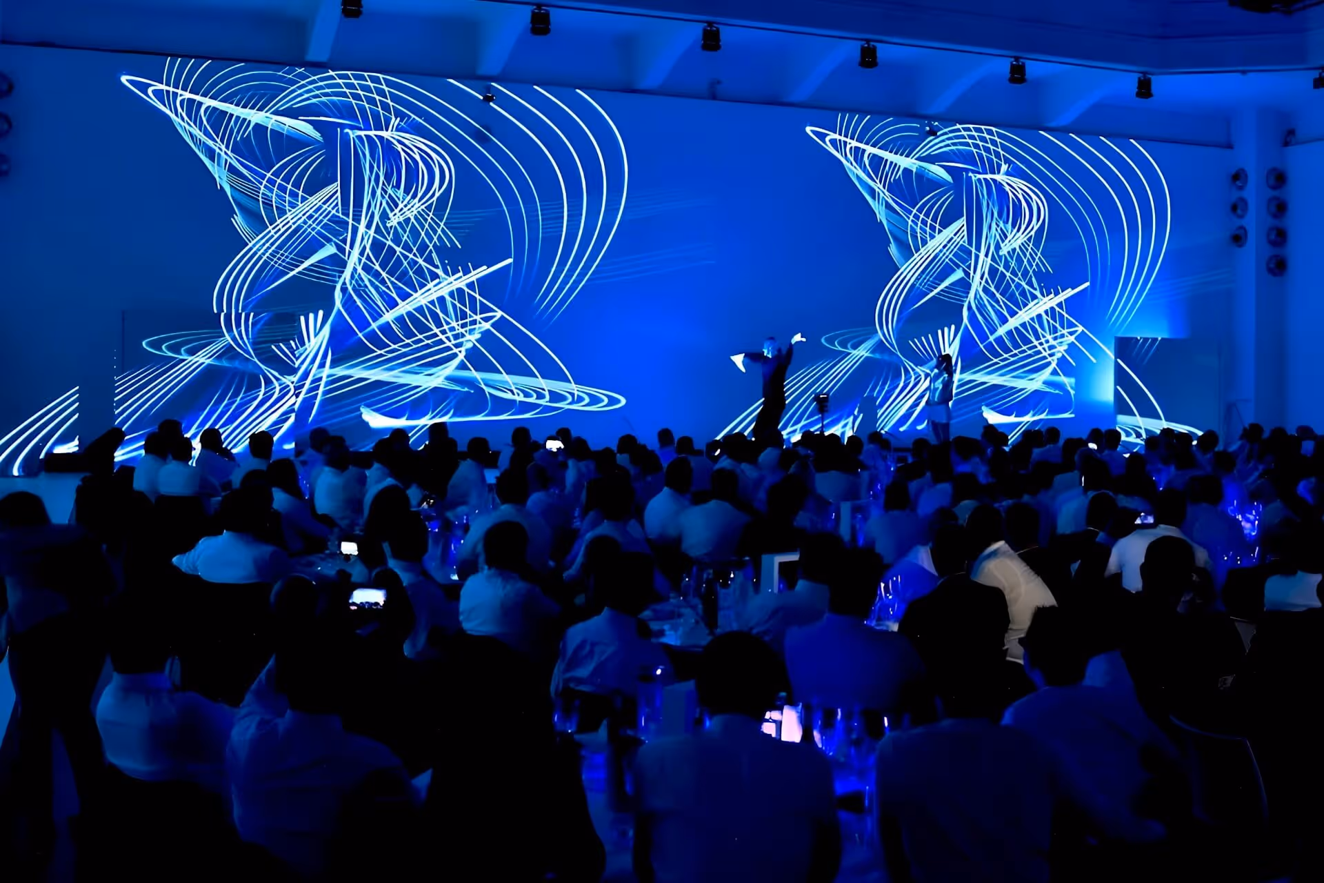 Publikum sitzt im Dunkeln vor einer Bühne mit blauen, geschwungenen Lichtmustern und Performern in einer Lichtshow.