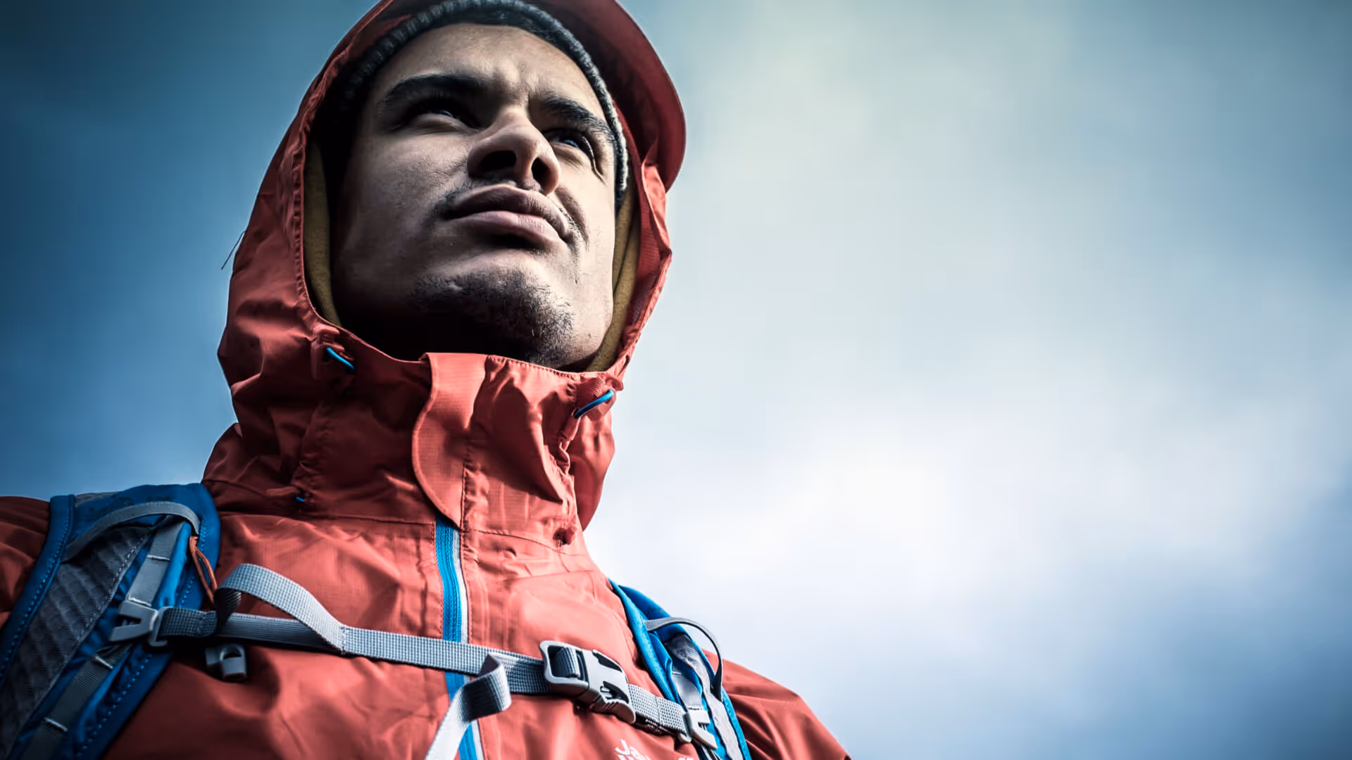 Mann mit roter Outdoorjacke und Rucksack blickt entschlossen in den Himmel bei bewölktem Wetter.