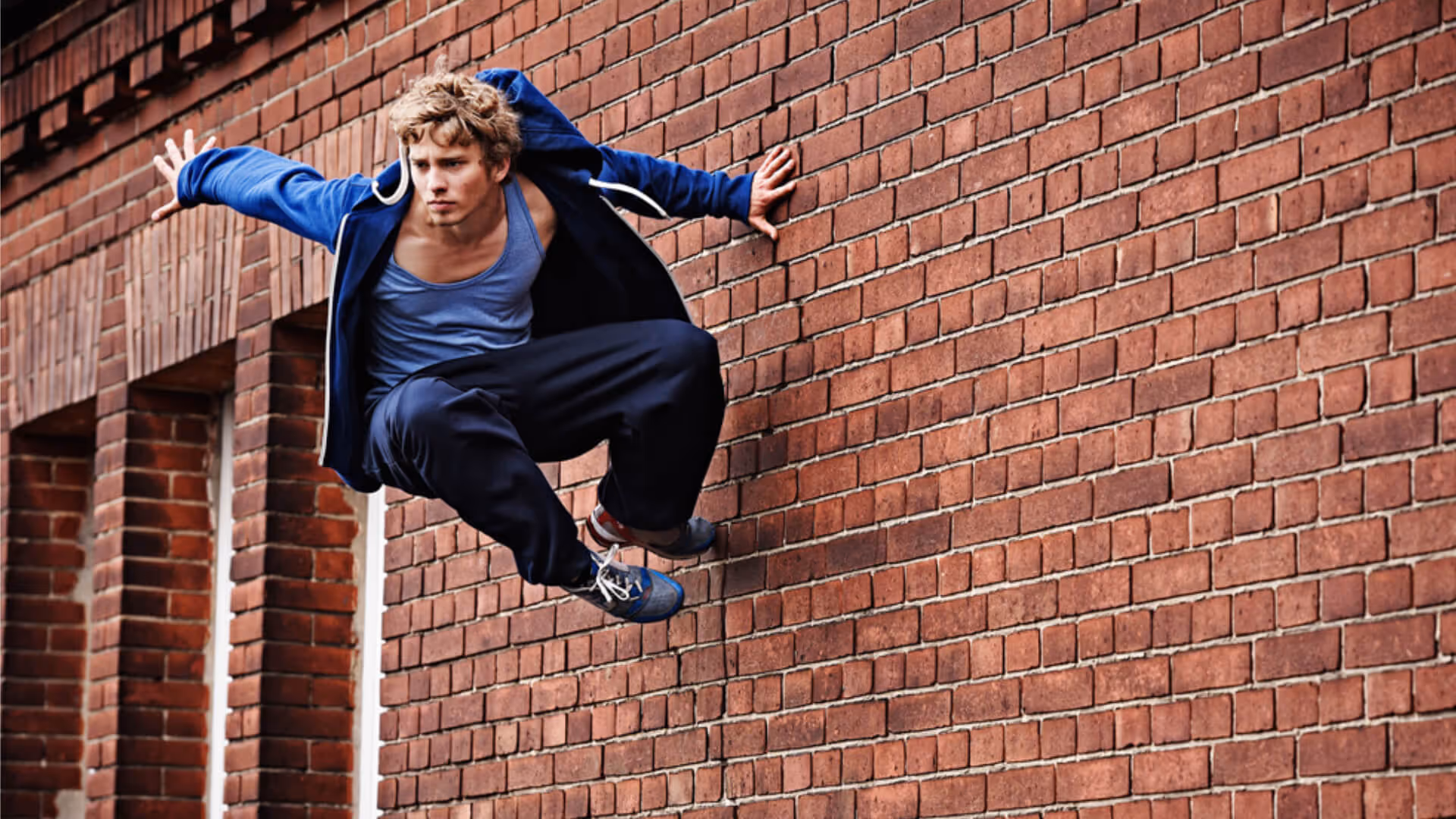 Junger Mann in blauer Kapuzenjacke und Sportkleidung, der dynamisch eine rote Backsteinmauer beim Parkour hochspringt.