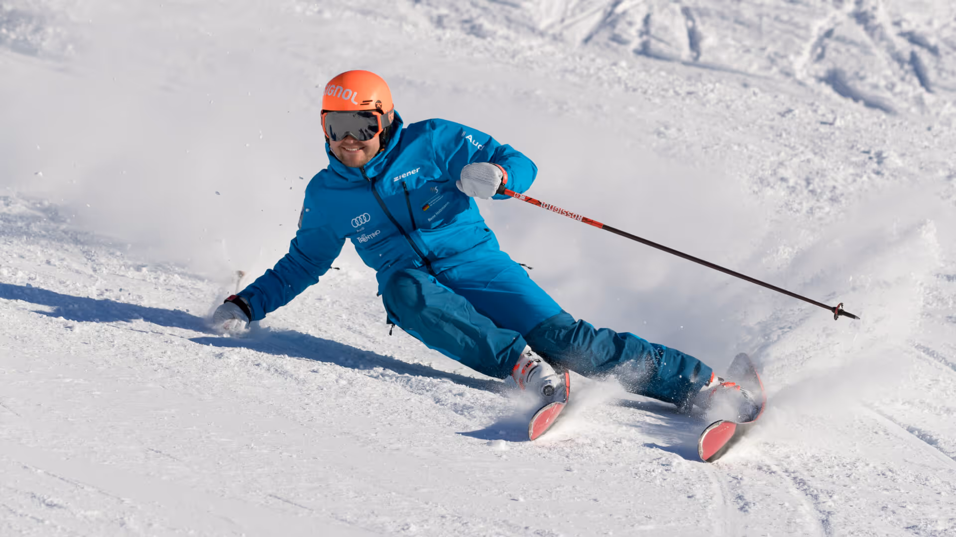 Ein Skifahrer in blauer Kleidung und rotem Helm fährt dynamisch eine schneebedeckte Piste hinunter und wirbelt Schnee auf.