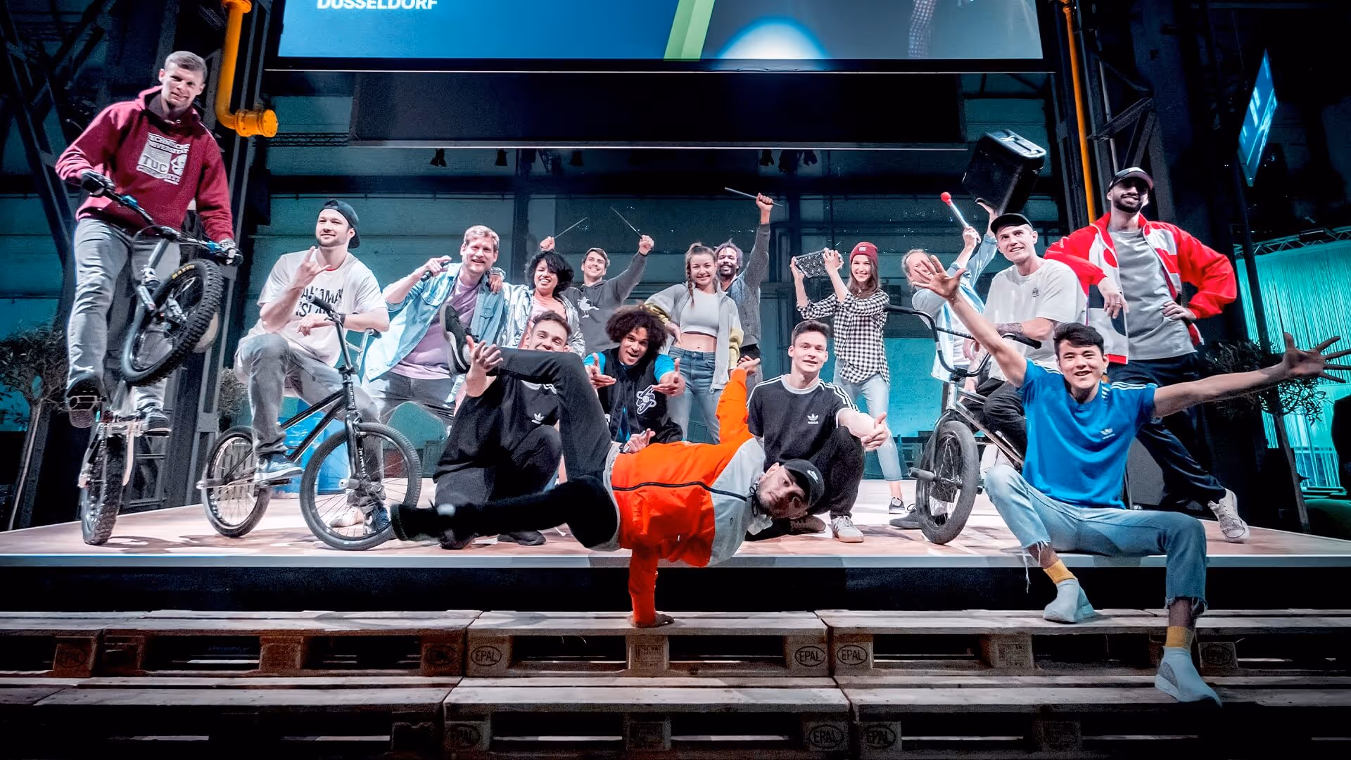 Eine Gruppe von jungen Menschen posiert fröhlich auf einer Bühne mit BMX-Rädern, Breakdance und Schlagzeugstöcken.