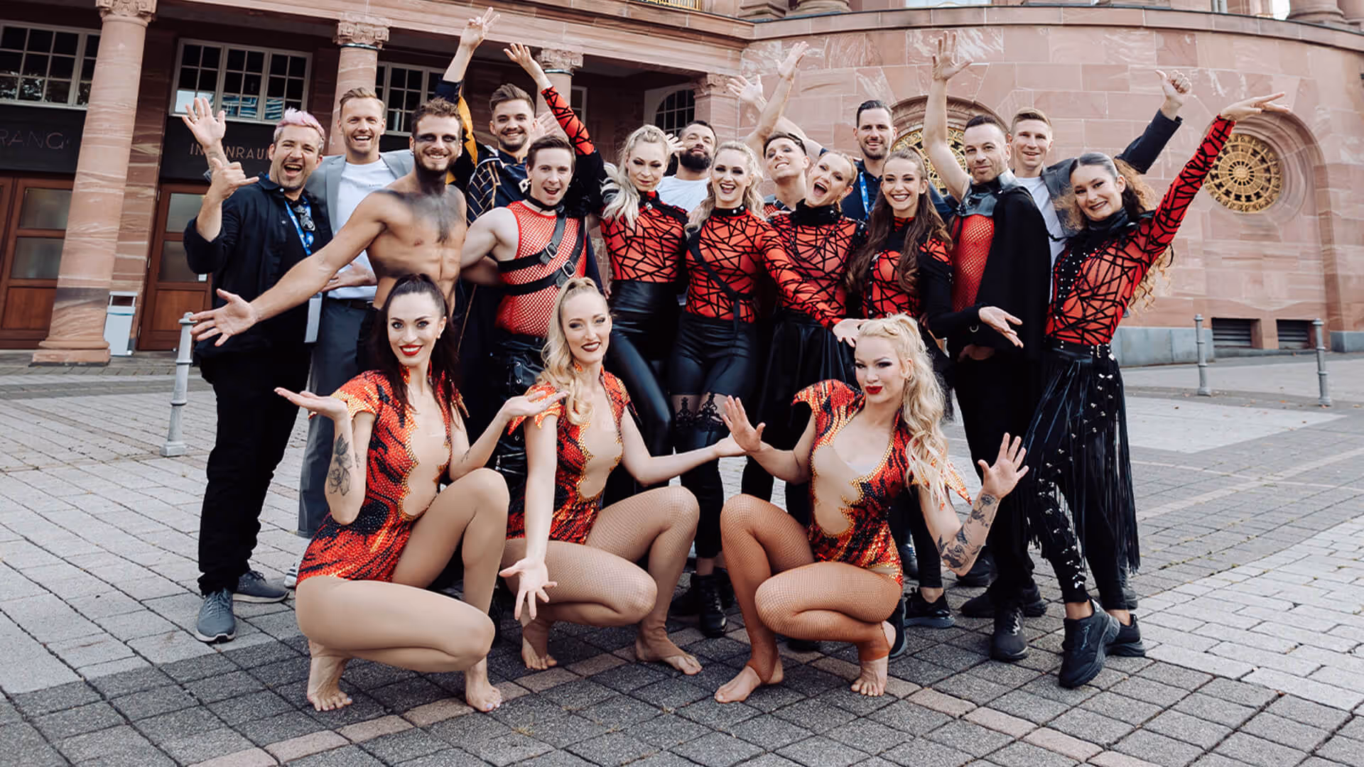 Gruppe fröhlicher Tänzer und Künstler posiert vor einem historischen Gebäude, viele tragen rote und schwarze Kostüme.