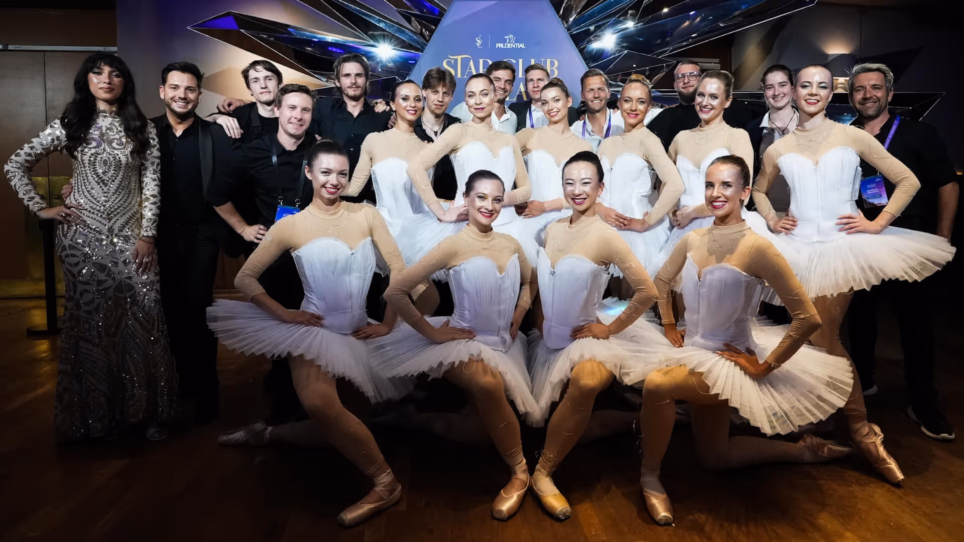 Gruppe von Balletttänzerinnen in weißen Tutus und Begleitpersonen vor dunklem Hintergrund bei einer Veranstaltung.