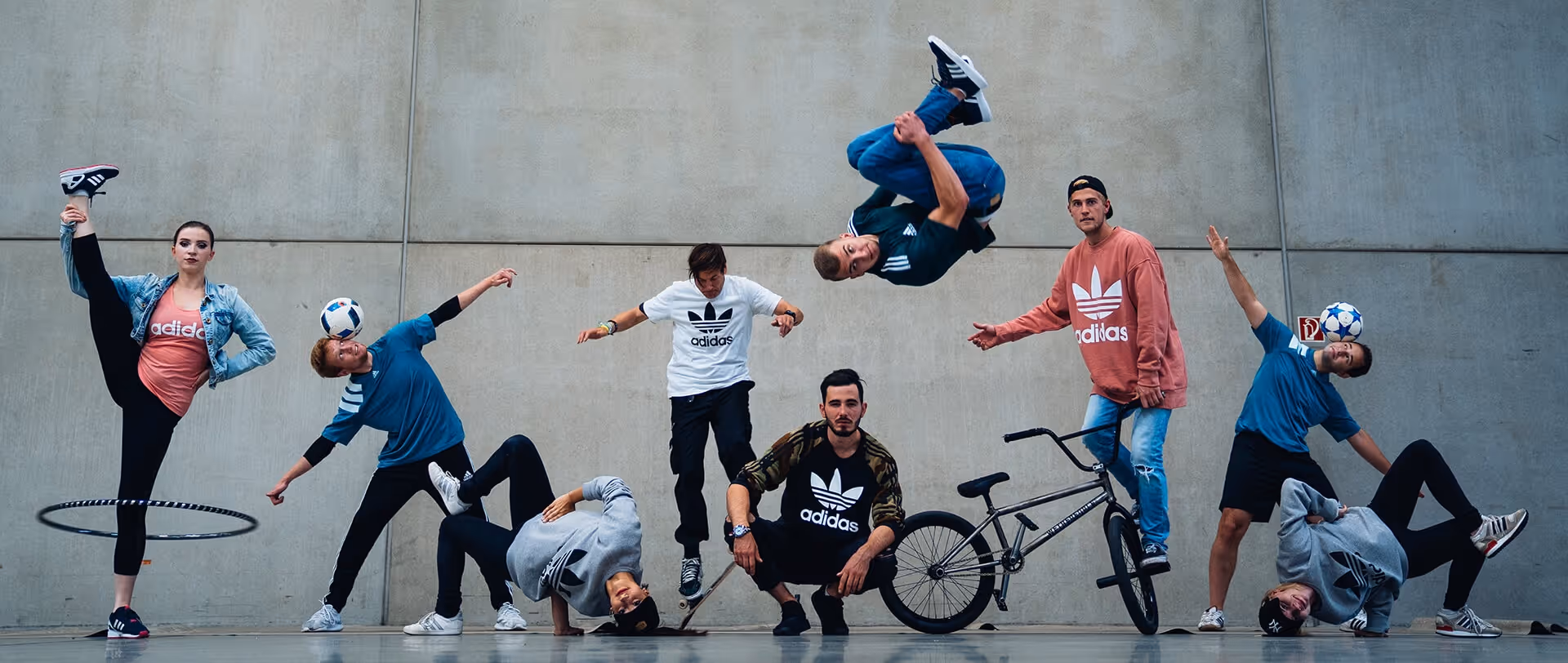 Acht junge Menschen tragen Adidas-Kleidung und zeigen verschiedene urbane Sportarten und Tricks vor einer grauen Wand, darunter Hula-Hoop, Fußballjonglage, Skateboarding, BMX und Breakdance.