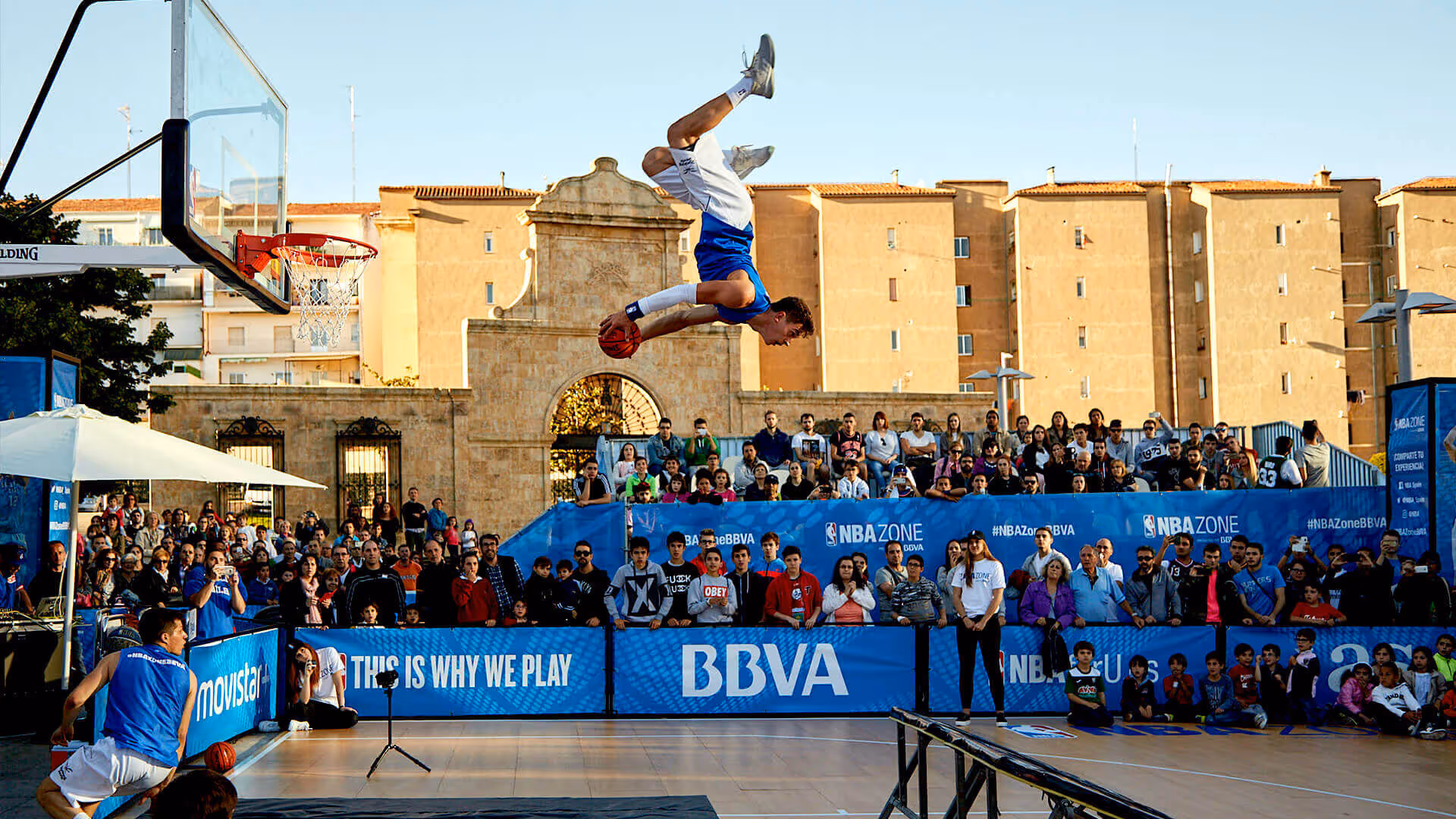 Ein Basketballspieler führt einen Luftsalto mit Ball vor einer begeisterten Menschenmenge bei einem Outdoor-Event durch.
