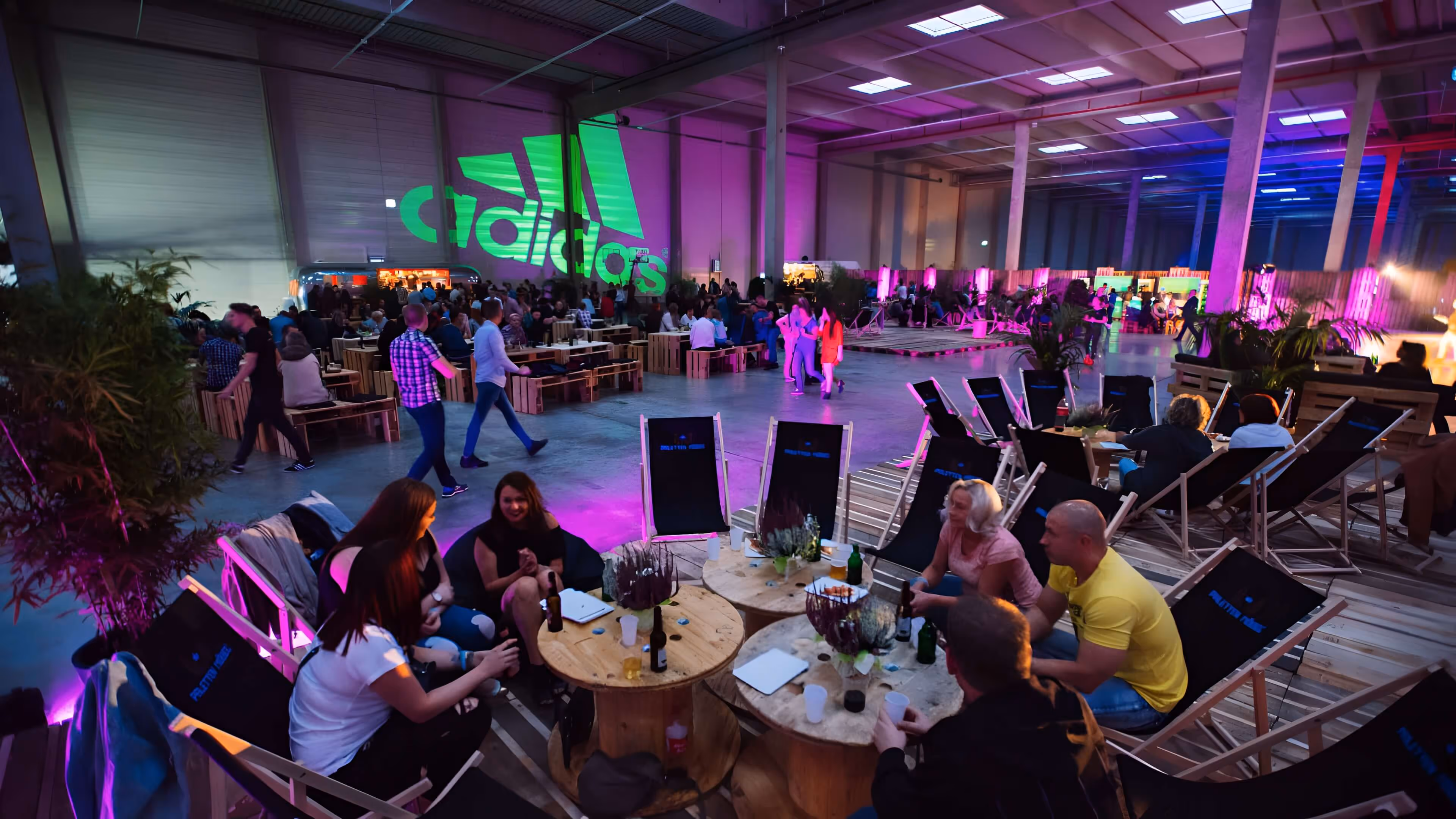 Große Veranstaltung in Lagerhalle mit Leuten, die an Tischen und in Liegestühlen sitzen, beleuchtet von bunten Lichtern und großem grünem Adidas-Logo an der Wand.