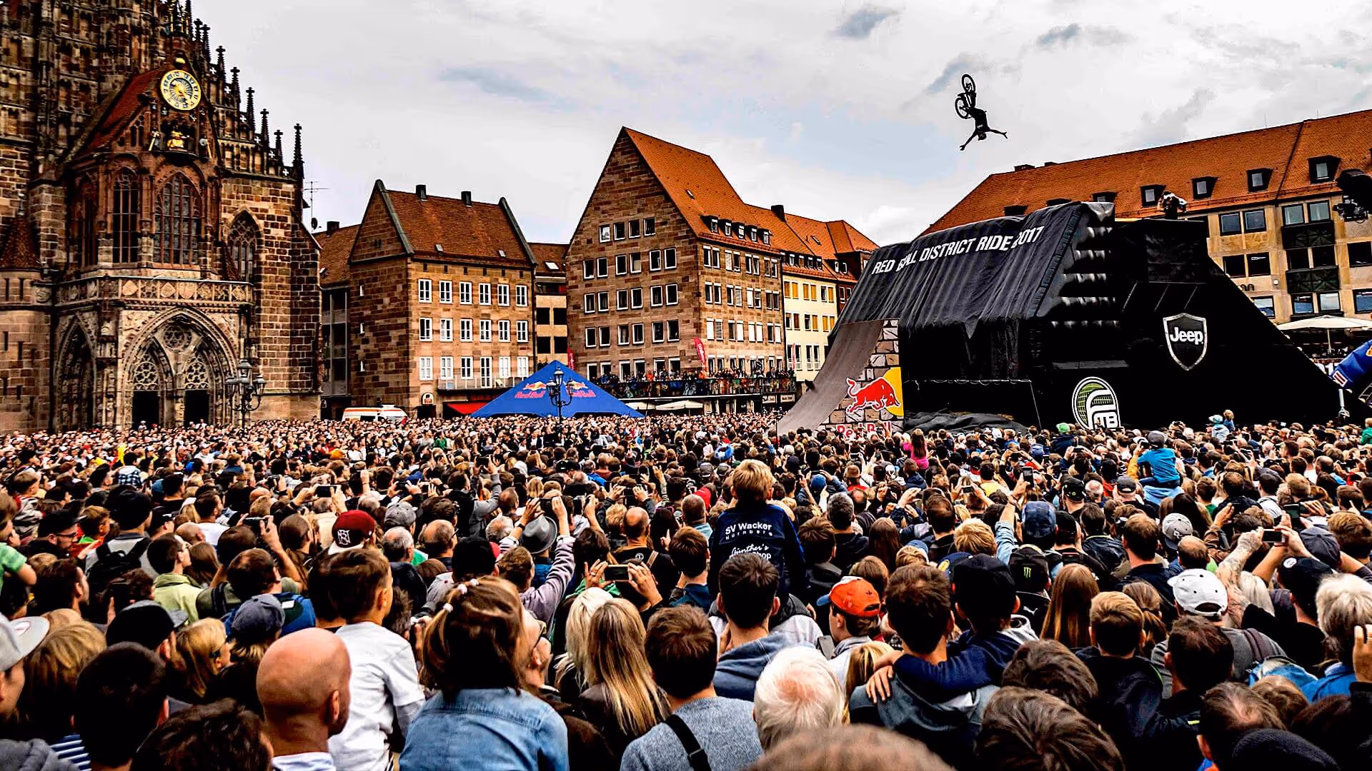 Große Menschenmenge vor historischen Gebäuden beobachtet einen BMX-Fahrer bei einem Sprung während eines Outdoor-Events.