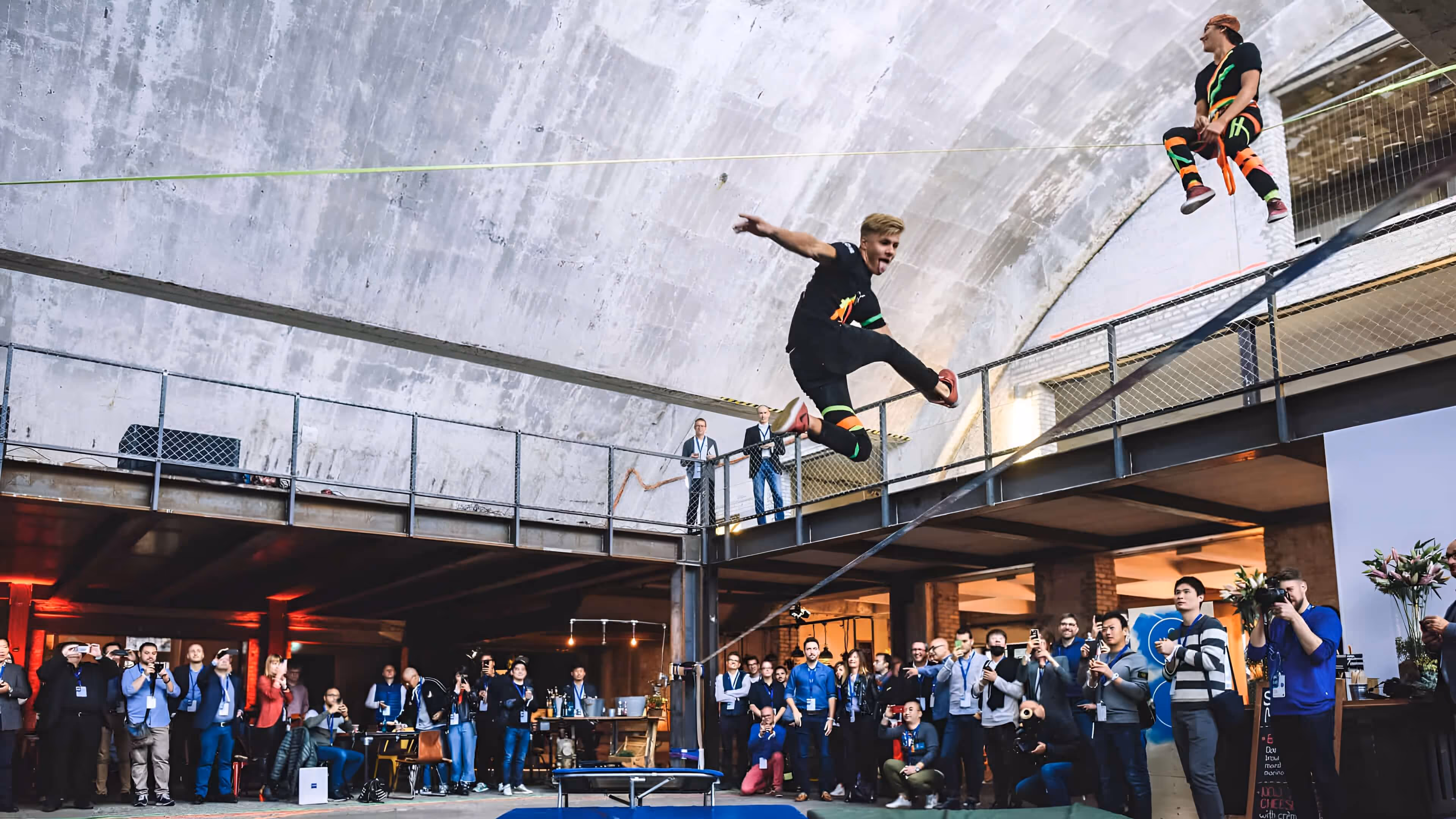 Zwei Männer in schwarzer Sportkleidung führen im Gebäude einen Slackline-Sprung über eine Gruppe zuschauender Menschen aus.