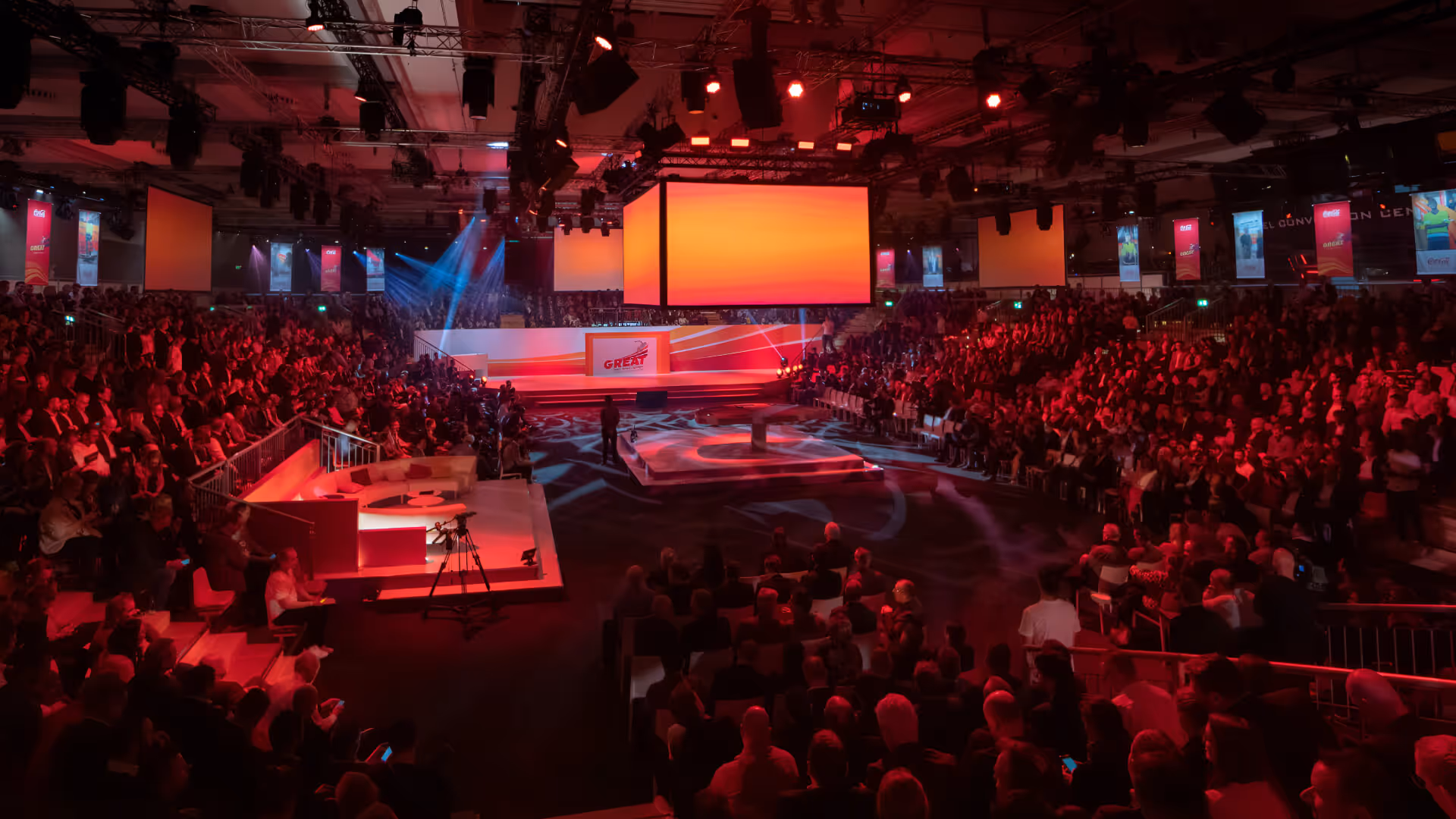 Große, vollbesetzte Veranstaltungshalle mit roter Bühnenbeleuchtung und einem zentralen Podium unter einem großformatigen, leuchtenden Bildschirm.