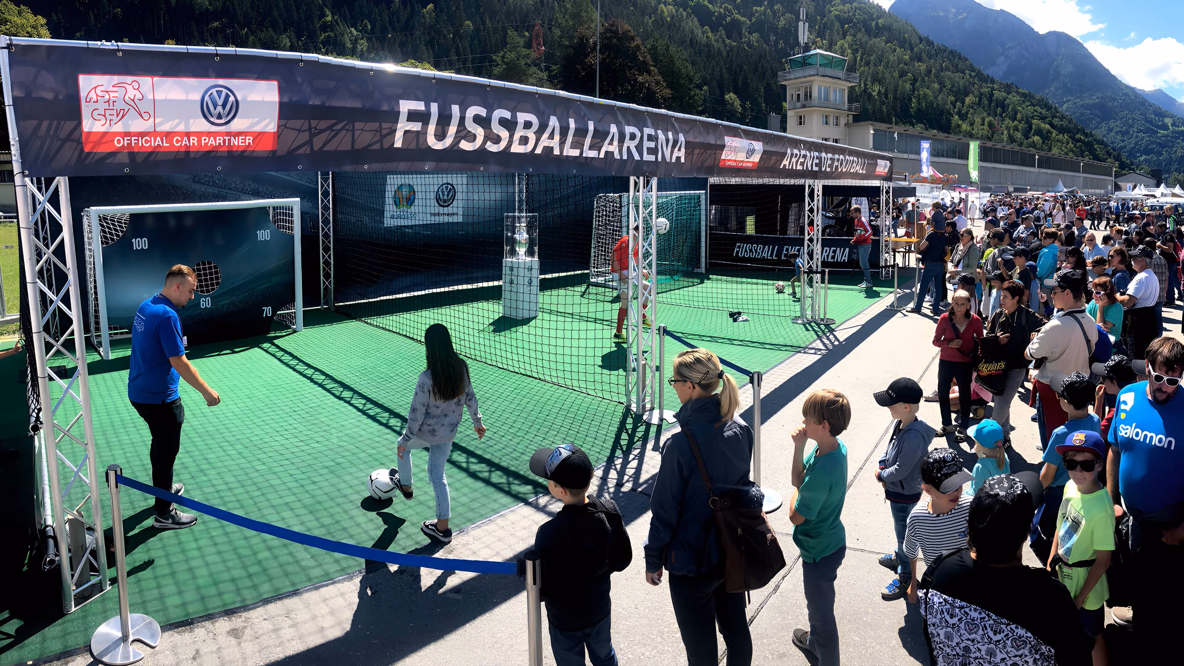 Menschen stehen in einer Schlange vor einem Fußballspiel-Eventbereich mit grüner Spielfläche und Torwandschießen, umgeben von Bergen.