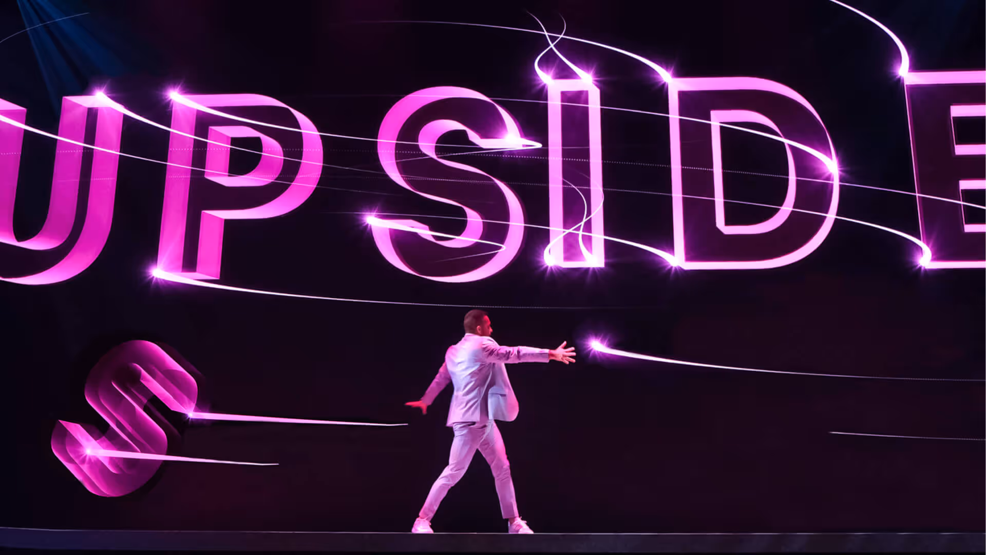 Ein Mann in einem weißen Anzug steht auf einer Bühne vor einer großen, leuchtenden pinken Schrift mit Lichteffekten.